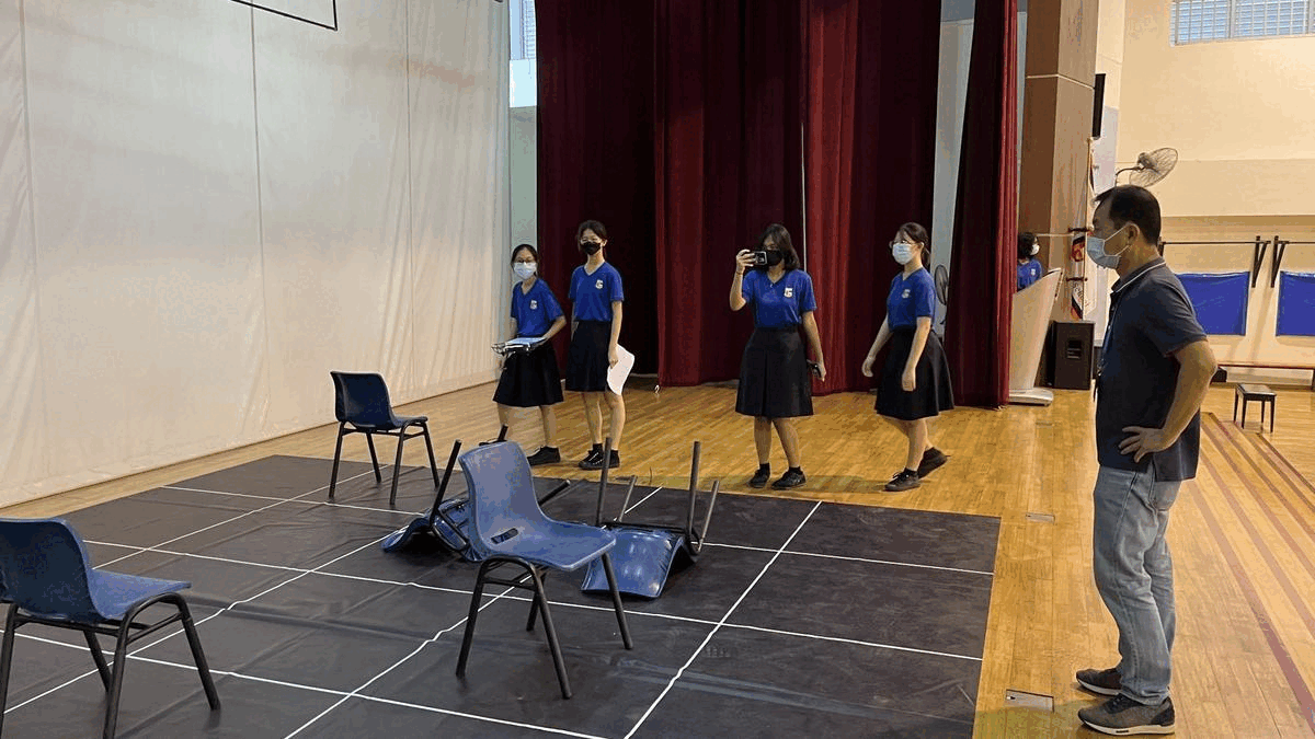 Four masked students in blue uniforms stand on a stage mat facing an adult man, with overturned chairs nearby.
